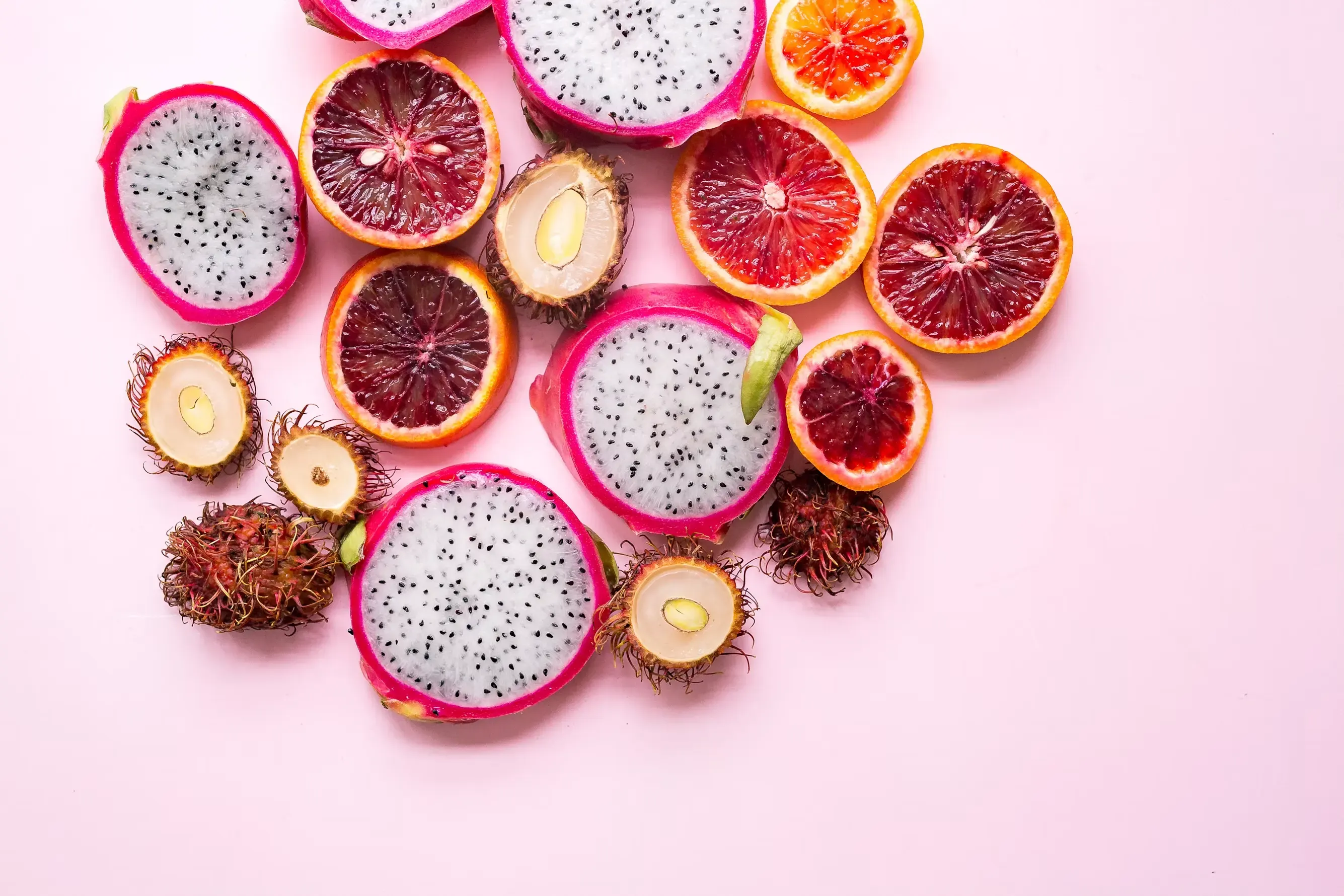 Blood oranges, dragon fruit and rambutans cut in half on a pink background, seen from above.