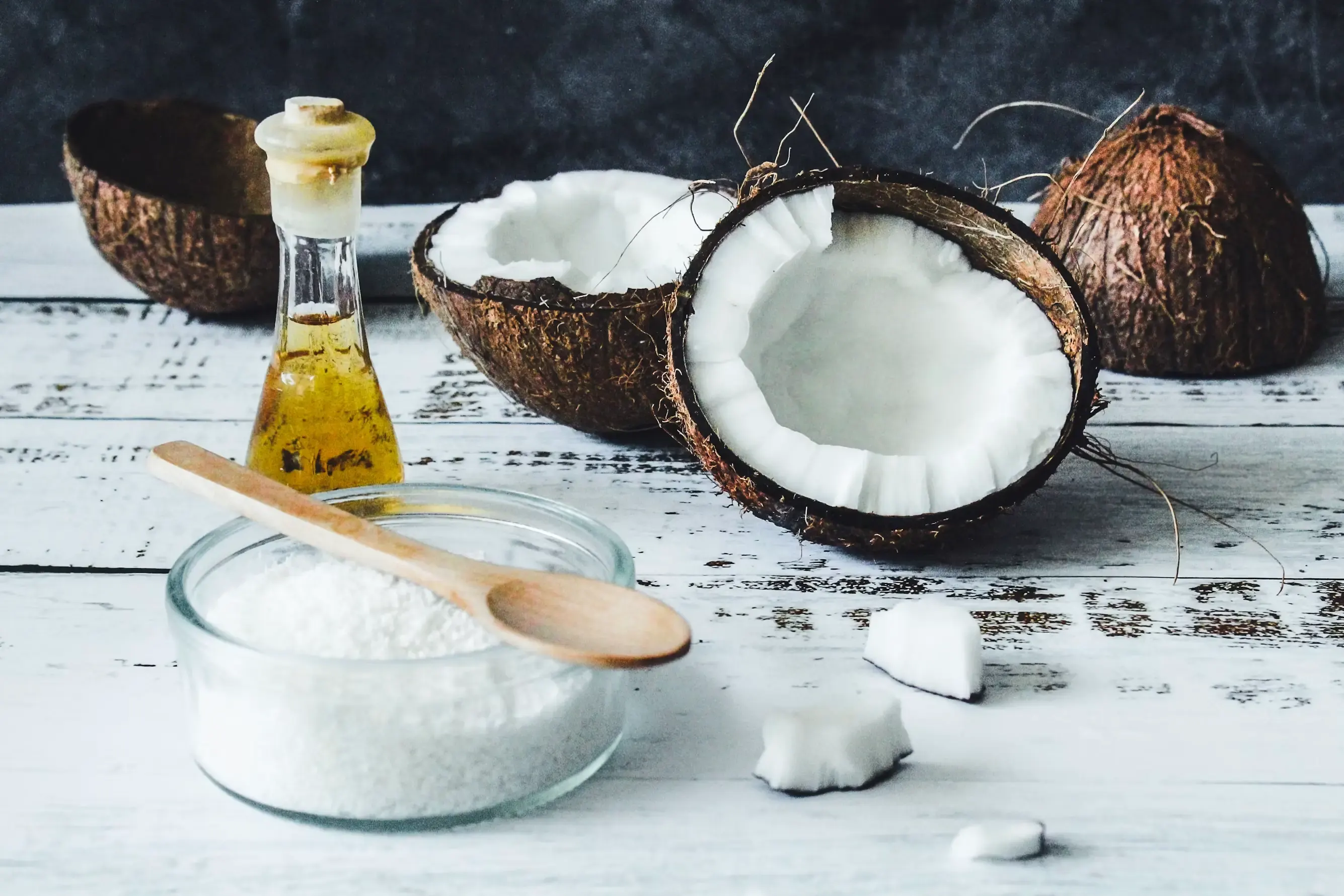A coconut split in two, a bottle of coconut oil and a bowl of grated coconut on a white wooden table.