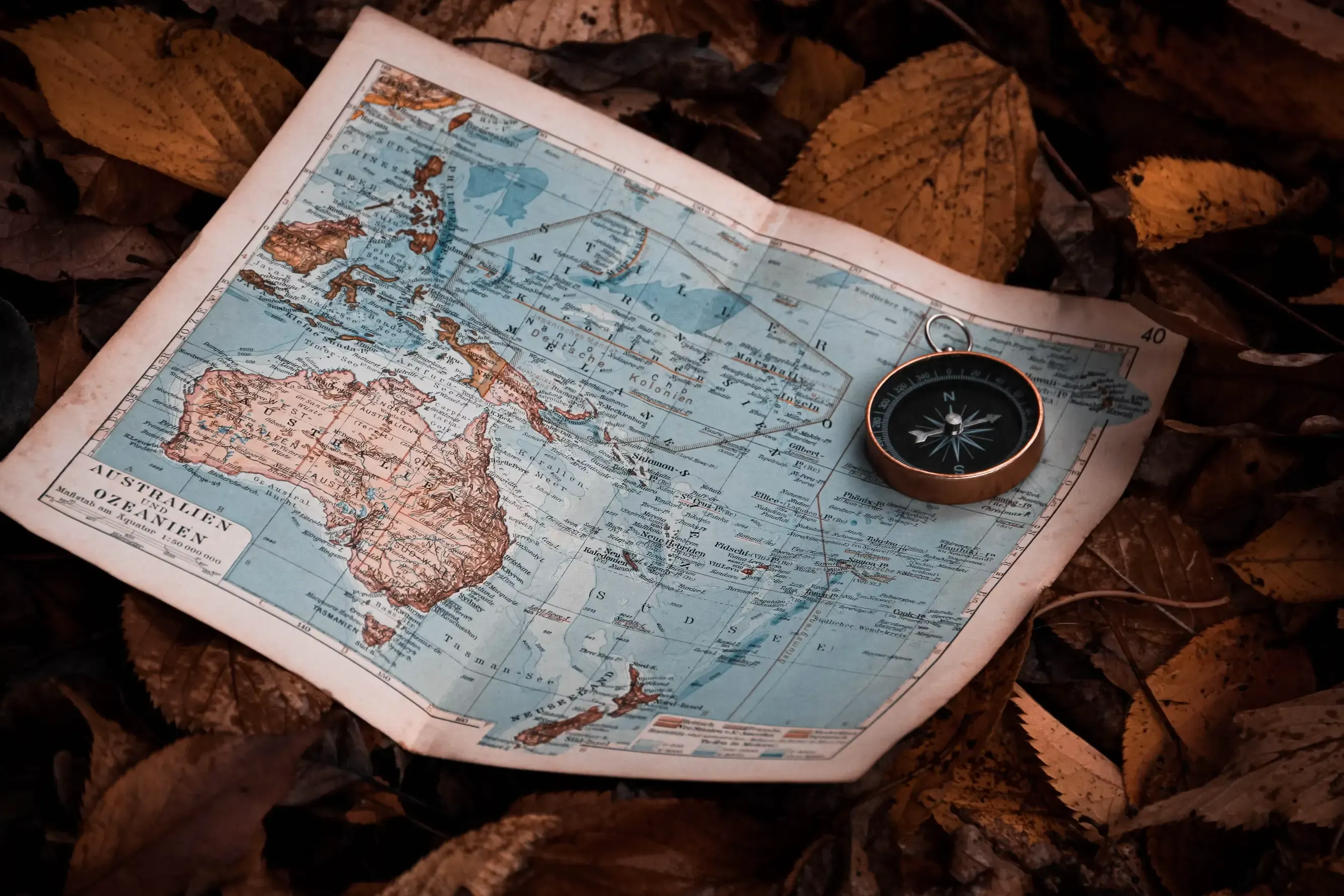 A brass compass resting on an old map of Australia and Oceania spread out over dry autumn leaves.