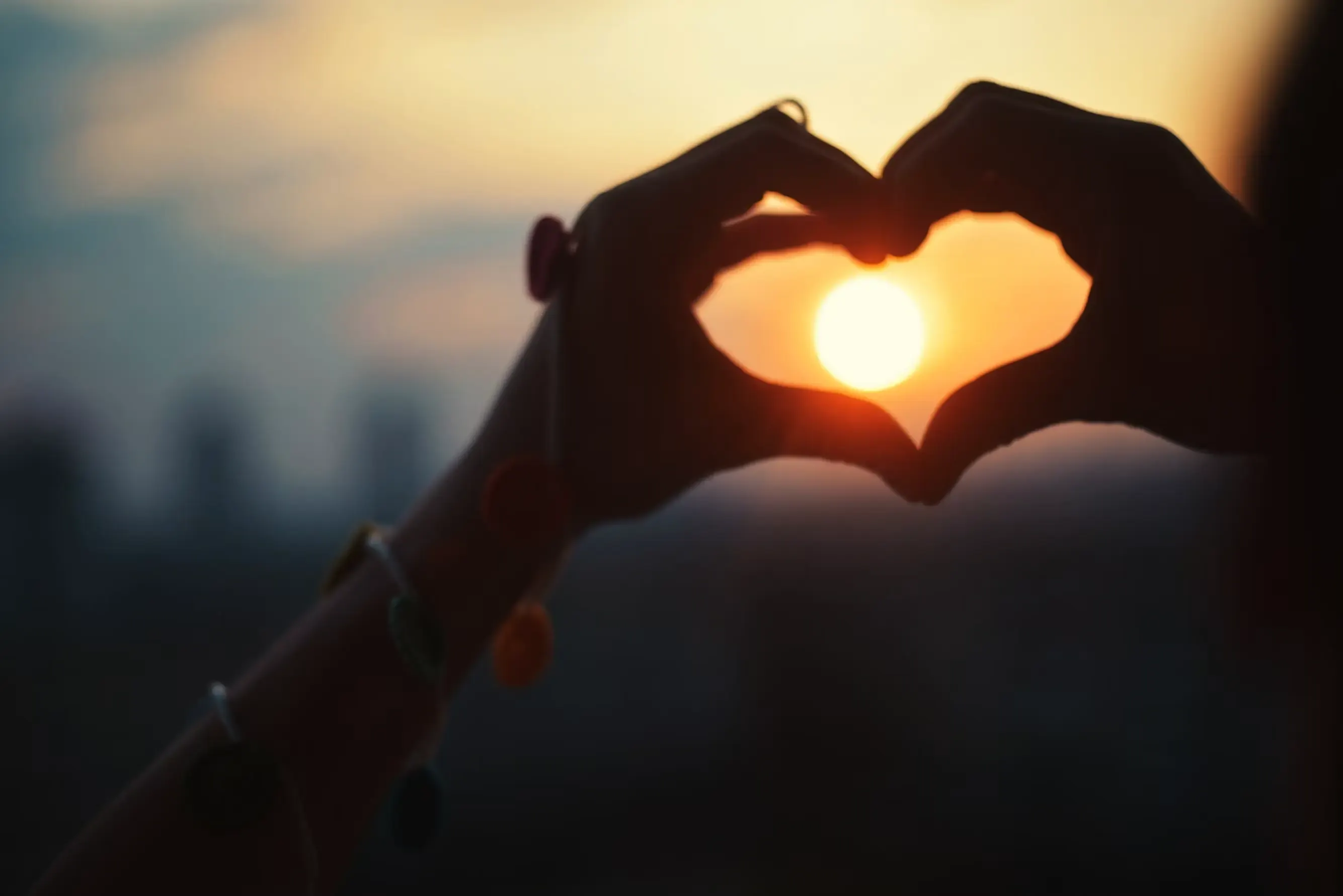 Two hands in silhouette form a heart against the light, framing the sun inside it at dusk.