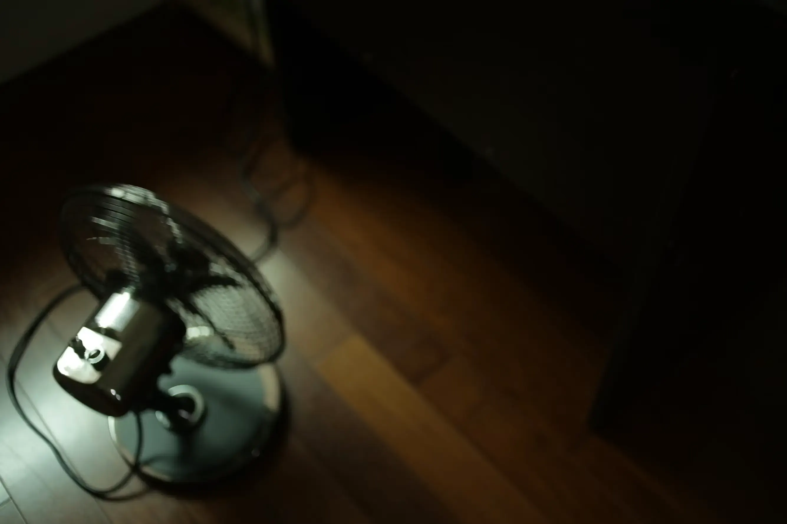A black and grey desk fan standing on a brown wooden parquet floor.