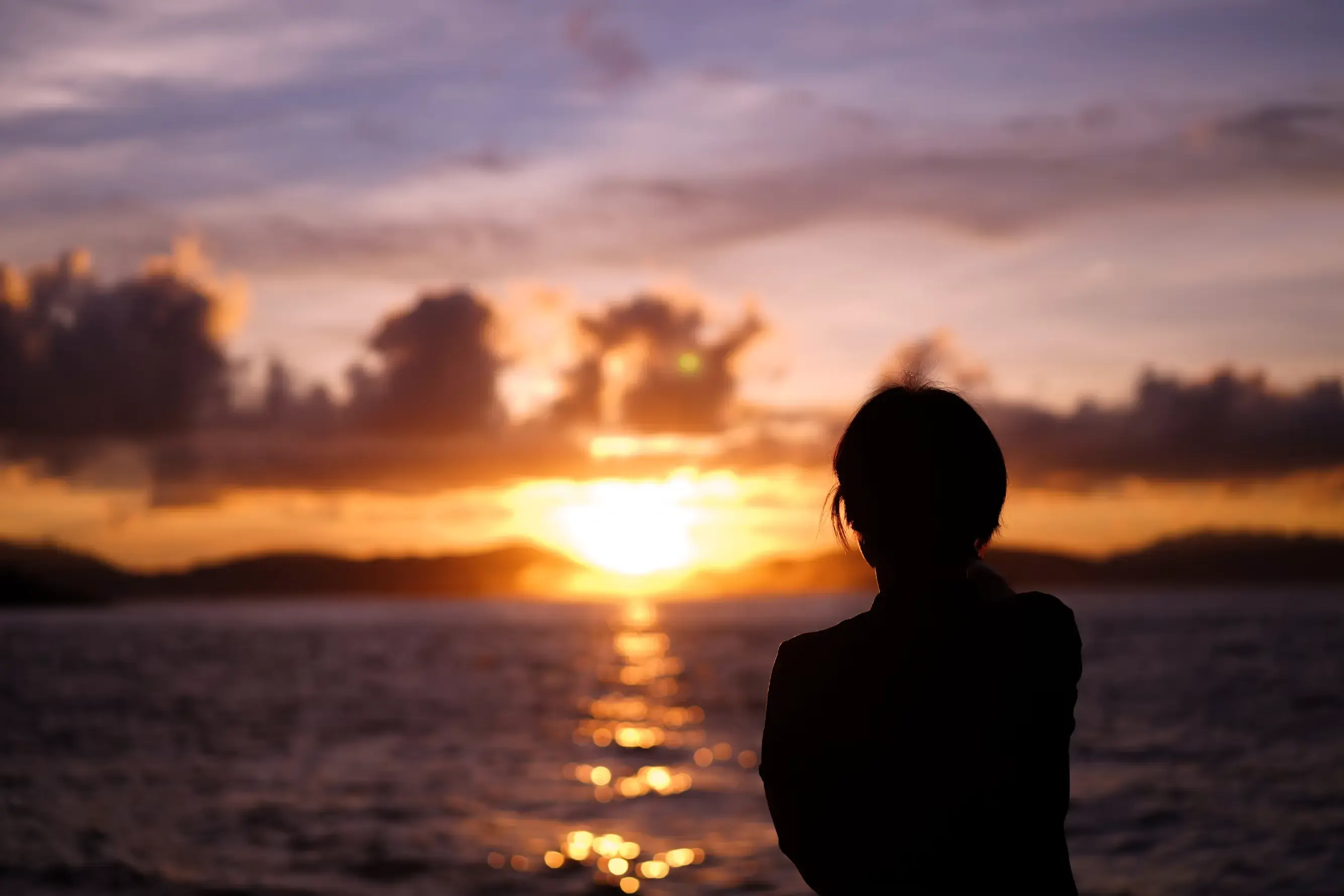 Backlit silhouette of a person seen from behind, in the foreground on the right, watching the sun set over the sea among orange clouds; the light traces a golden trail across the water.