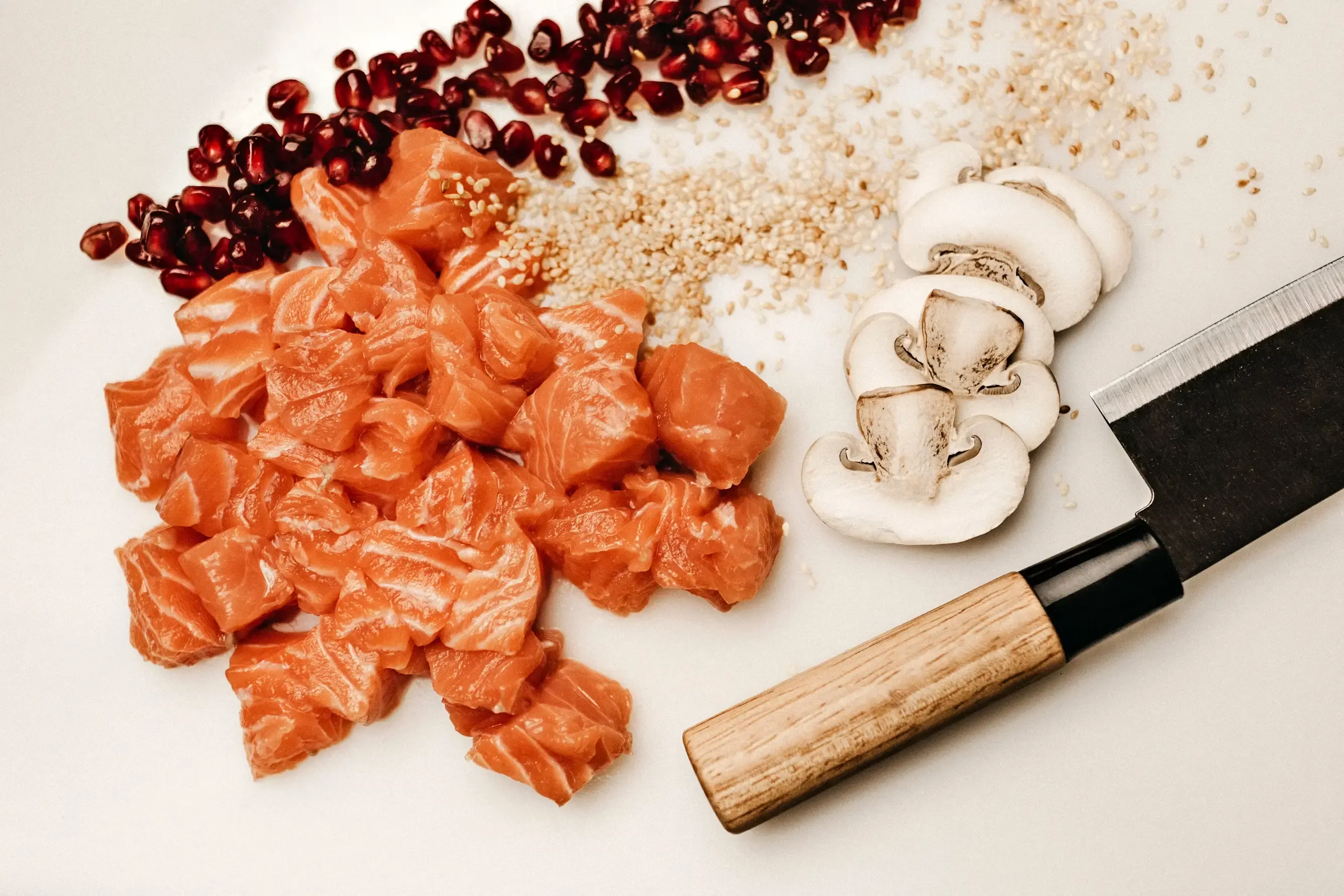 Overhead view of a white surface with raw salmon cut into cubes, three slices of white button mushroom, scattered sesame seeds and pomegranate seeds in the top left corner. On the right, a Japanese knife with a dark blade and a pale wooden handle.