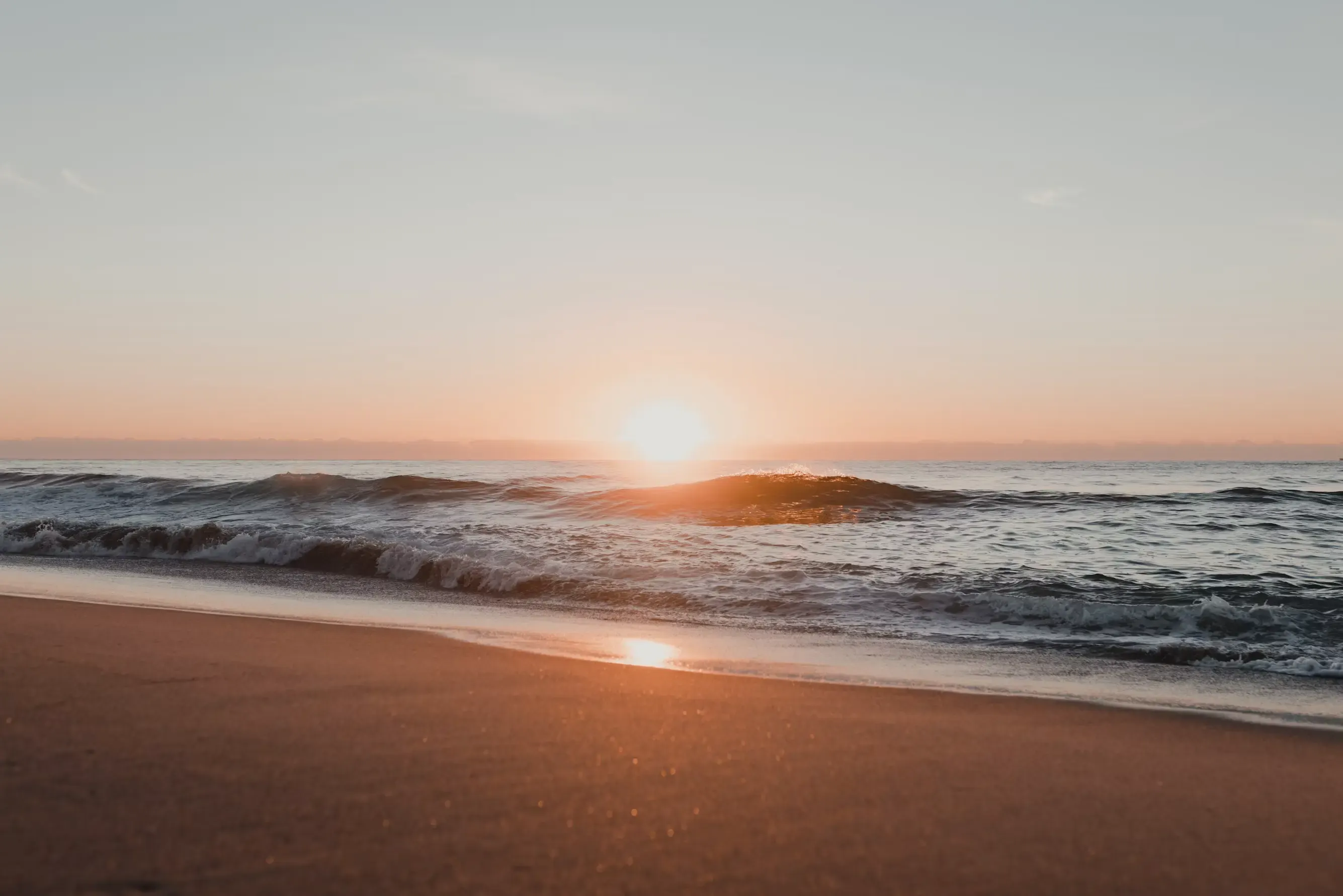 The sun rising on the horizon over the sea, with a wave breaking on the shore and its light reflected on the wet sand of the beach.