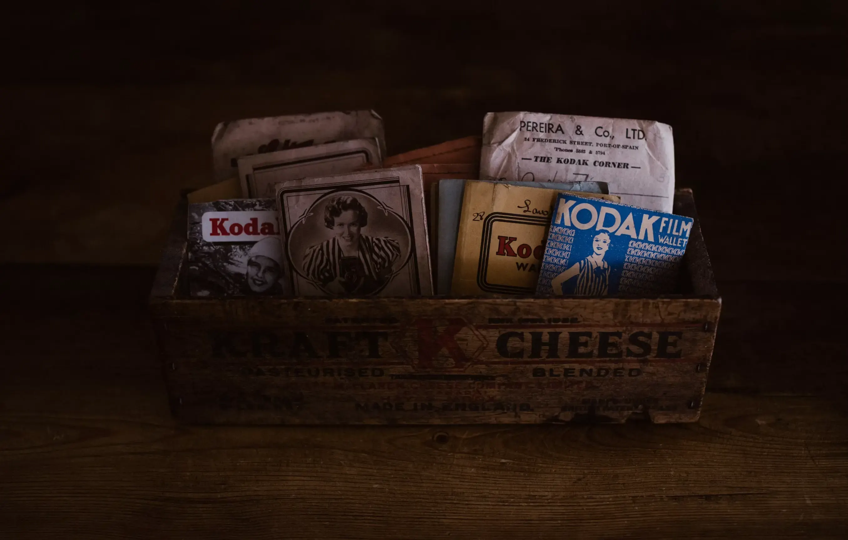 A wooden crate stamped «Kraft Cheese», filled with old Kodak film wallets and sepia portraits, on a wooden table in dim light.